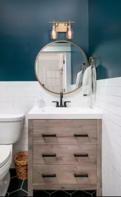 A bathroom with a light wooden vanity with a white sink and black faucet. Above it is a round mirror and above is a gold light fixture. The top half of the walls are painted a dark blue and the bottom half have white subway titles.