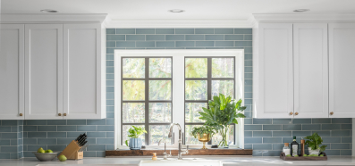A kitchen with a blue subway tile backsplash a large window and white cabinets.
