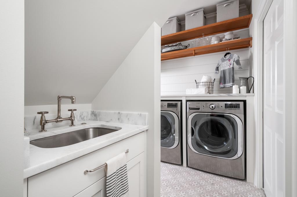 A white laundry room with sink, and wooden shelves above a washer and dryer