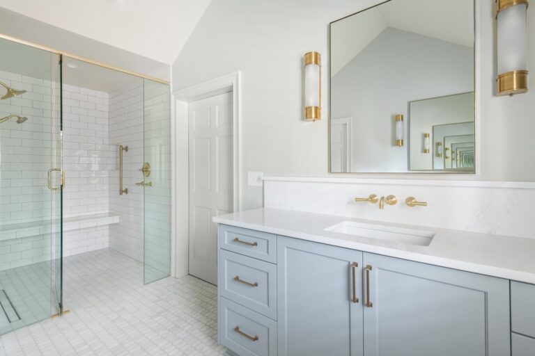An all white bathroom, with gold accents, a shower, and a large vanity with a white sink and counter top, and powder blue cabinets.