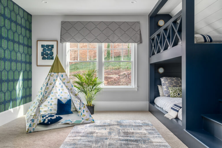 A children's bedroom with a dark blue and green accent wall, a teepee and rug on the floor, and a large window with fabric shades. Opposite the accent wall is a navy blue built-in bunk bed with stairs.
