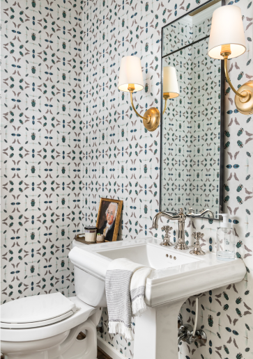 A powder room with white, green, and gray abstract patterned wallpaper, with a toilet next to a white sink under a tall mirror with 2 gold sconces on either side.