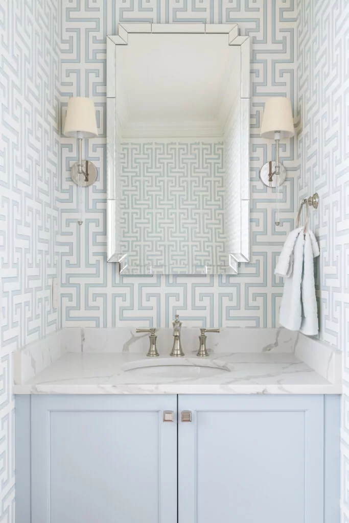 A powder room with white and blue printed wallpaper, a large mirror with 2 sconces on either side. The sink has marble countertops and powder blue cabinets underneath.