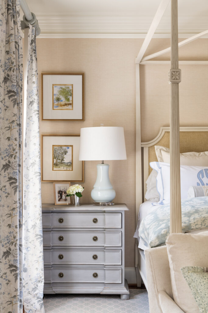 A corner of a beige, white and pastel bluee bedroom with a large bed-side table and floral curtains next to a bed.